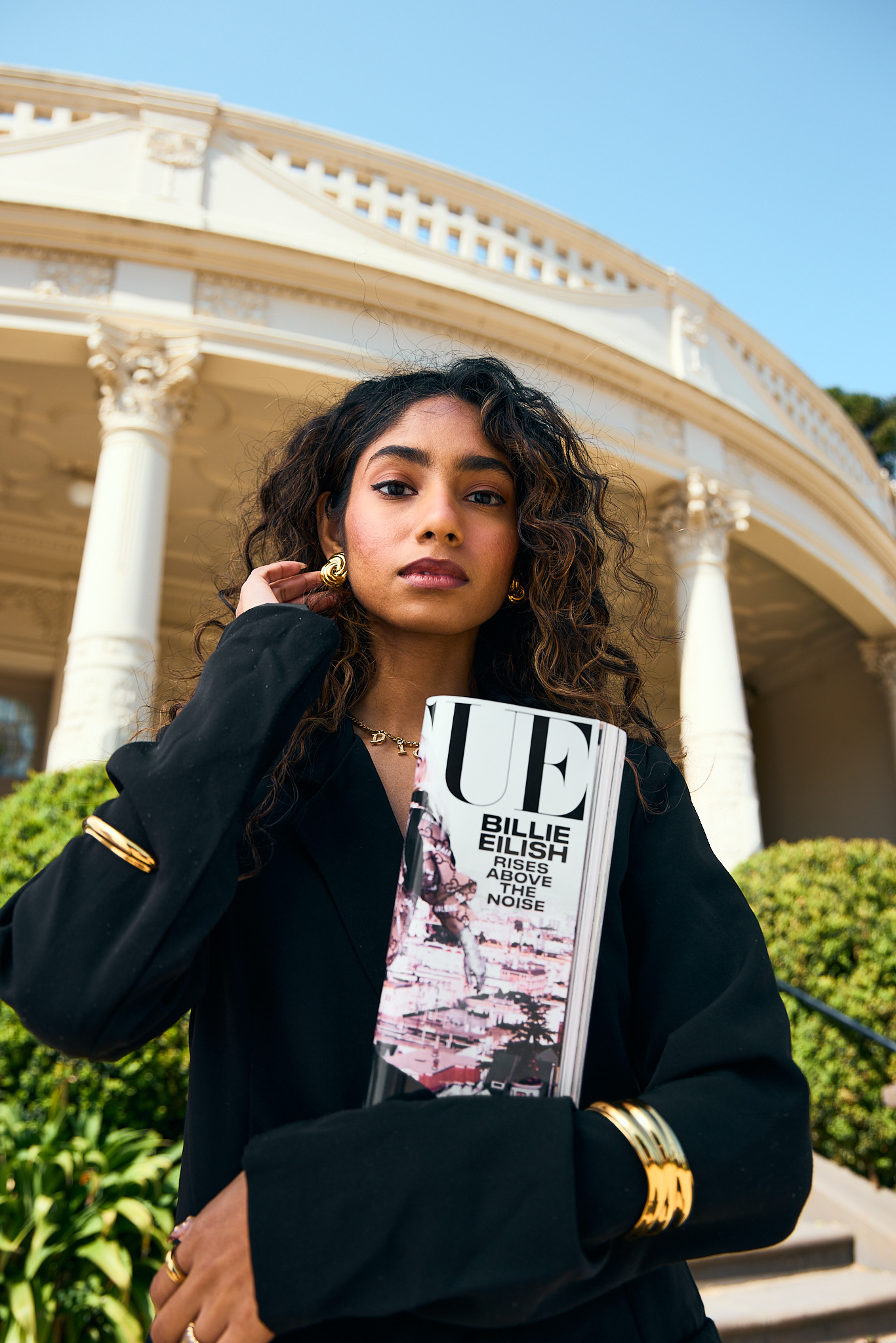 Young woman poses outdoors, holding Vogue magazine with Billie Eilish cover against a classical building with columns and bright sky.